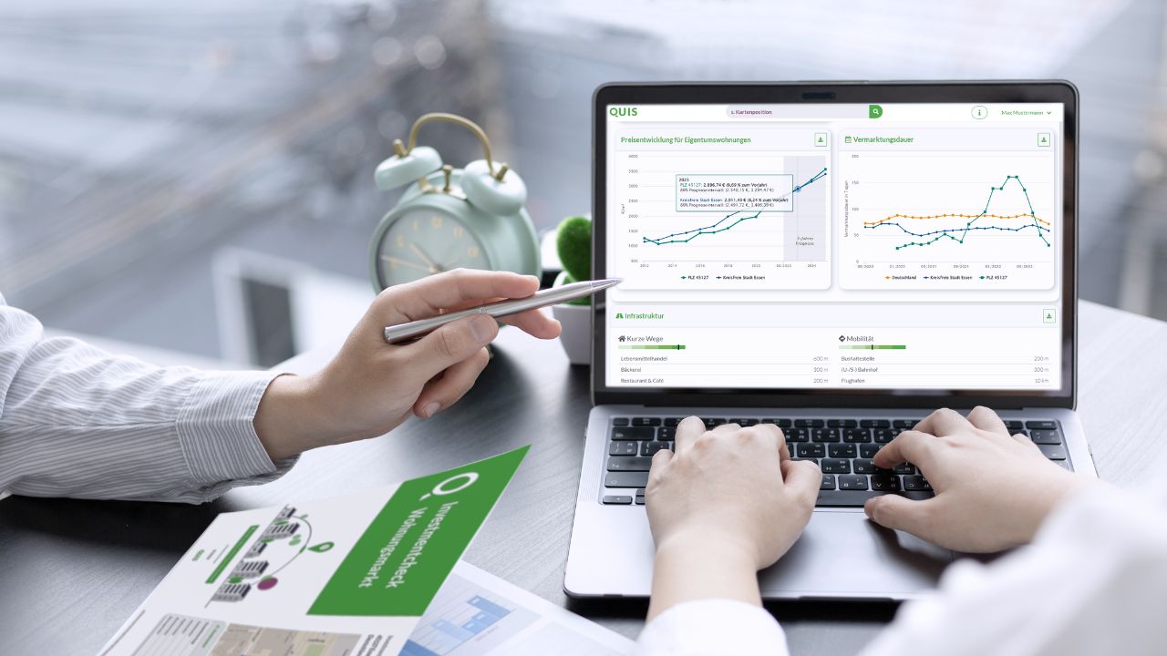 Makler in einem Büro analysiert Immobilienmarkt-Daten auf einem Laptopbildschirm, der Diagramme zur Preisentwicklung und Vermarktungsdauer zeigt. Ein grüner Flyer liegt auf dem Tisch, und ein grüner Wecker steht daneben.