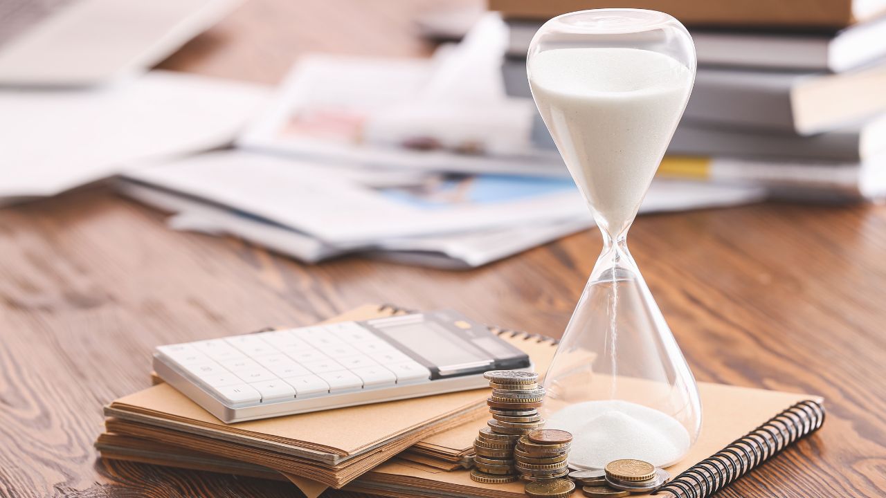 Ein Stundenglas auf einem Holztisch, daneben gestapelte Münzen, ein Taschenrechner und Notizbücher, im Hintergrund unscharfe Papierstapel, symbolisch für Zeitmanagement und Finanzen.