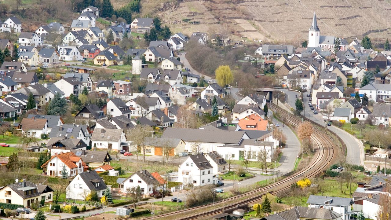 Luftaufnahme einer dicht bebauten Ortschaft mit zahlreichen Ein- und Mehrfamilienhäusern, einer Kirche im Hintergrund und einer sich durch das Bild schlängelnden Eisenbahnlinie